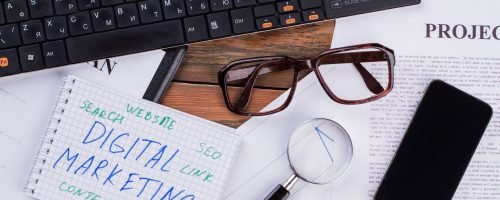 Digital marketing on notepad and various business papers on brown background. Brown glasses magnifier and smartphone near notepad.